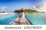Back view of a elegant woman in white dress and hat walks down a pier towards a tropical island in the Maldives
