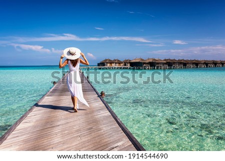 Back view of a elegant woman in white dress and hat walks down a pier over turquoise ocean in the Maldives islands