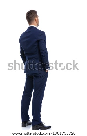 back view of elegant young businessman in navy blue suit holding hands in pockets and standing isolated on white background in studio