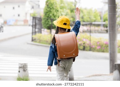 The back view of an elementary school student wearing a school backpack and raising their hand to cross at a crosswalk; an image illustrating traffic safety for students on their way to school