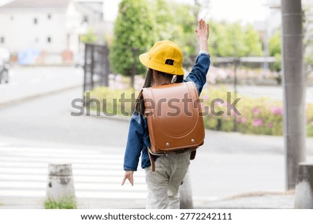 The back view of an elementary school student wearing a school backpack and raising their hand to cross at a crosswalk; an image illustrating traffic safety for students on their way to school