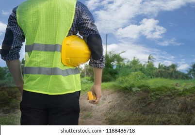 Back View Of Engineer, Architect, Supervisor Worker, Wearing Green Reflective Vest, Holding Yellow Safety Helmet, Water Level Gauge At Construction Site, Empty Land. Real Estate, Home Building Concept