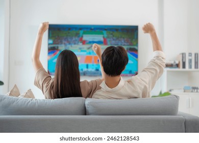 Back view of an enthusiastic couple sitting on a sofa, arms raised in victory while watching a live sports match on their home television, expressing excitement and support - Powered by Shutterstock