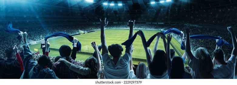 Back view of excited football, soccer fans cheering their team with blue scarfs at crowded stadium at evening time. Concept of sport, cup, world, team, event, competition
