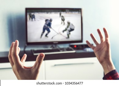 Back View Of Excitement Man With Gesturing Hands Up How Watching Ice Hockey In Television At Home. Rear View Of Male Watching Sport In TV. An Excited Fan Is Rooting For His Favorite Team