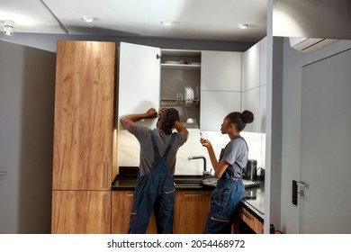 Back view experienced handyman fixing cupboard door in modern kitchen. Young lazy apprentice passively looking at working partner. African american service workers in contemporary kitchen interior. - Powered by Shutterstock