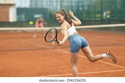 Back view of expressive focused young female tennis player engrossed in game on clay court, swinging racket ready to hit ball over net - Powered by Shutterstock