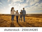 Back view of family of four hugging each other and looking into the distance enjoying sunset and beautiful nature standing in a row in the field together. Young parents with children walking outdoors