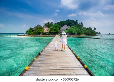 Back view family mother and daughter in white dress walking on wooden deck near the beach enjoying tropical summer vacation. Background water bungalow and the beauty of the sea with the coral reefs
