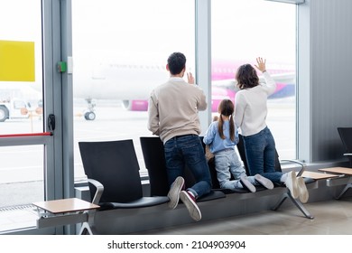 Back View Of Family Waving Hands While Looking At Plane Through Window In Airport