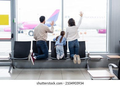 Back View Of Family Waving Hands While Looking At Airplane Through Window In Airport