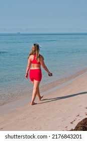 Back View, Far Distance Of A Young, Adult Female In A Two Piece, Orange Bathing Suit, Walking A Tropical Shoreline, On A Sunny Morning