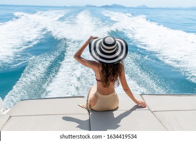 Back view: Fashion photo of adorable young woman sitting on edge of luxury yacht and looking for the sea during sailing trip. Happy woman enjoying summer travel.