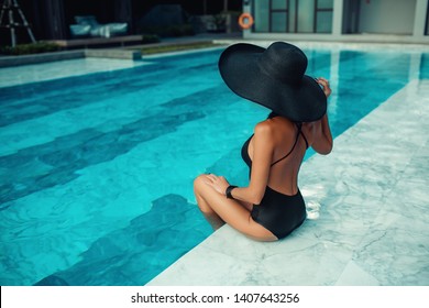 Back view: Fashion stylish young woman posing while sitting near the swimming pool. Hat, swimsuit, Outdoor fashion, elegant black hat