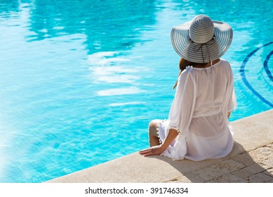 Back View Of Fashion Woman On Summer Vacation Relaxing At Luxury Resort Spa Poolside. Young  Fashionable Lady Wearing Sun Hat And White Kaftan.