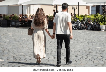 Back view of fashionable young couple in love walking in the town square holding hands. Rear view of romantic young married couple spending time together outdoors. City street life. Relationship - Powered by Shutterstock