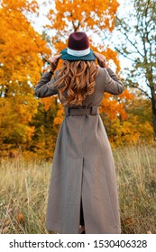 Back View Of A Fashionable Young Woman In A Stylish Long Coat In An Elegant Hat With A Trendy Hairstyle In An Autumn Park Among Trees With Orange Foliage. Modern Girl Walks Through The Woods.