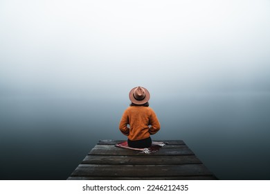 Back view of fashioned young woman sitting on wooden dock looking at view on a misty morning. Female hipster with brown hat relaxes on the edge of jetty admiring foggy lake. Wonderful nature getaway - Powered by Shutterstock