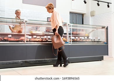 Back view of fasionable lady looking and choosing fresh meat in store. Cheerful senior male butcher in apron standing behind store counter with big assortment of raw meat products. - Powered by Shutterstock