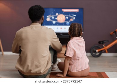 Back view of father and daughter watching educational videos about space together sitting on floor in front TV - Powered by Shutterstock