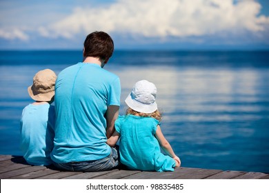 Back View Of Father And Kids Sitting On Wooden Dock Looking To Ocean