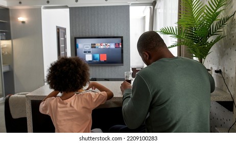 Back View Of Father And Little Son Having Lunch Or Dinner With Cola And Pizza At Table At Home Kitchen. Unhealthy Eating. Young Black Family Lifestyle And Relationship. Fatherhood And Parenting