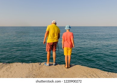 Back View Of Father And Son Fishing Together From Jetty. The Concept Of The Transfer Of Experience From The Older Generation