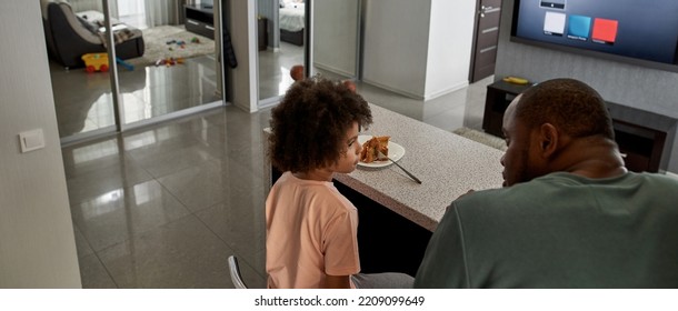 Back View Of Father And Son Looking At Each Other During Having Lunch Or Dinner With Pizza At Table At Home Kitchen. Unhealthy Eating. Black Family Lifestyle And Relationship. Fatherhood And Parenting