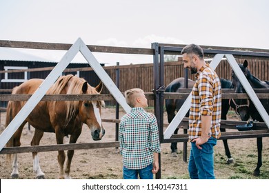 back view of father and son smiling each other while standing near horses at ranch