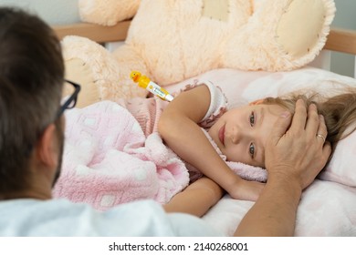 Back View Of Father Touches Forehead Of Sick Little Child With Fever And Measure Temperature With Thermometer. Treatment For Illness, Flu And Cold