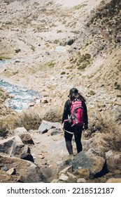 Back View Of Female Hiker Walking Through A Rocky Path Beside A Mountain River, With A Backpack On. Vertical Shot