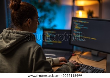 Back view of female programmer using laptop and pc while working at home during evening time. Concept of coding, freelance and IT sphere.