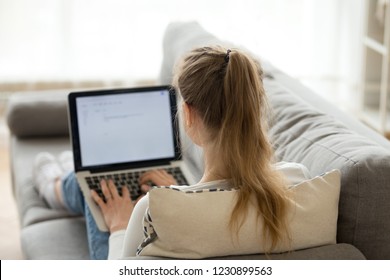 Back view of female relax on cozy couch with laptop on knees texting or chatting with friends, girl lying on comfy sofa writing email working from home, student studying at computer resting in bed