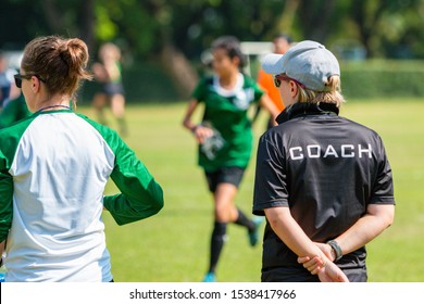 Back view of a female soccer, football, coach in black coach shirt standing in the sun watching her team play at an outdoor football field