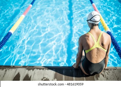 Back View Of Female Swimmer Who Sits On The Side Of The Swimming Pool. Woman Wears A Black-lime Swimsuit, A White Swim Cap And Swim Glasses. She Looks In Front Of Herself. Outdoors. Horizontal.