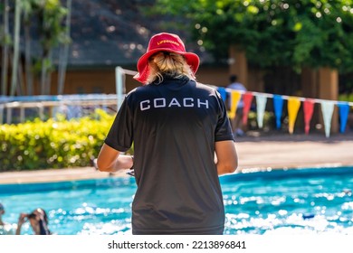 Back view of female swimming coach, wearing COACH shirt and red LIFEGUARD hat, working at an outdoor swimming pool - Shutterstock ID 2213896841