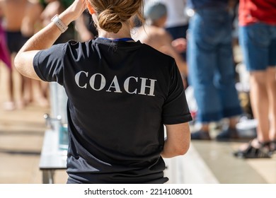 Back view of female swimming coach, wearing COACH shirt, working at an outdoor swimming pool - Shutterstock ID 2214108001
