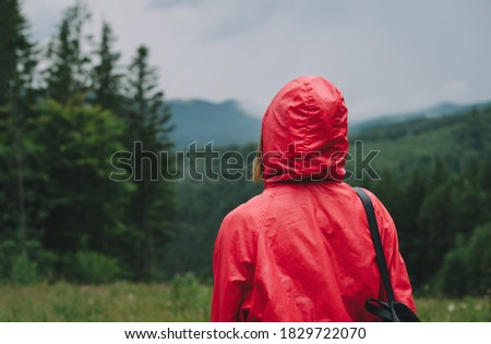 Back view of a female traveler in a red raincoat with a backpack looking at the mountain chain and picturesque view in the mountain fir forest.