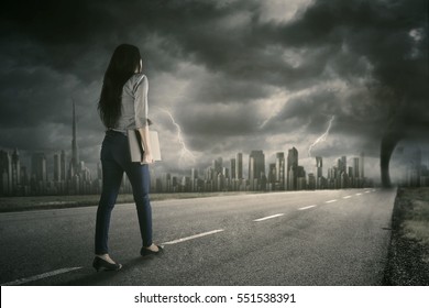 Back View Of A Female Worker Walking On The Street While Carrying A Laptop Computer With Tornado On The Sky