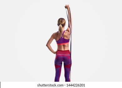 Back view of fit female in sportswear raising arm and pulling rubber band during training against white background