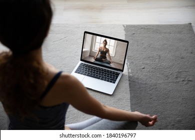 Back view of fit sporty woman sit on floor at home mediate practice yoga having video call with coach. Female do exercises watch stretching class on computer online, take distant course or lesson.
