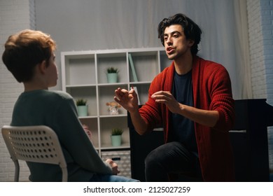 Back view of focused schoolboy sitting on chair listening young music teacher talking and gesturing at home during lesson. Music teacher explains to young student how to sing and play the piano. - Shutterstock ID 2125027259