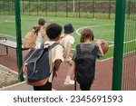 Back view of four schoolchildren entering sports ground or stadium with foorball field while one of them dribbling ball for playing basketball