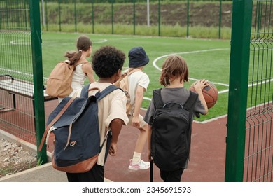 Back view of four schoolchildren entering sports ground or stadium with foorball field while one of them dribbling ball for playing basketball - Powered by Shutterstock