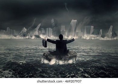 Back View Of Frustrated Businessman Sitting On The Rock At Sea While Looking At A Sinking City