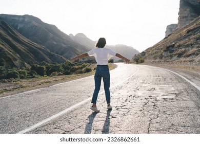 Back view full length of anonymous female tourist in summer wear with raised arms standing alone on empty road and enjoying freedom during trip through mountains