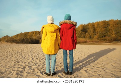 Back view of full length two young modern women friends standing together on sunny beach, stylish girlfriends on sea coast in colorful red yellow coat jacket and hat outdoors