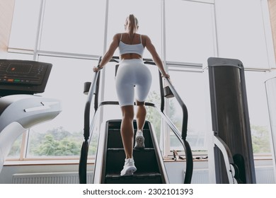 Back view full size young strong sporty athletic sportswoman woman in white sportswear warm up training running on a treadmill climber stairs machine in gym indoors. Workout sport motivation concept