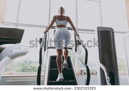 Back view full size young strong sporty athletic sportswoman woman in white sportswear warm up training running on a treadmill climber stairs machine in gym indoors. Workout sport motivation concept