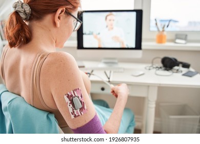 Back View Of Ginger Woman With Glucose Meter At Her Stomach Speaking On Video Call With Female Doctor, While Having Online Consultation At Home. Patient Engaged In Webcam Digital Conversation On